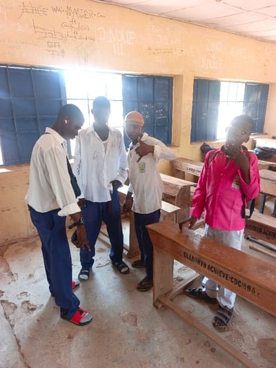 The scene depicts a classroom in Mada, Nigeria. Four young males, likely students, are present. The room features light-colored walls heavily covered with white graffiti and text, above two sets of barred windows admitting bright sunlight. The floor is rough, worn concrete. Several wooden desks or benches are visible, one in the foreground marked with "CLAP/HVN ACHIEVE/CDC/0AAI".

On the left, three males are grouped. The leftmost wears a white long-sleeved shirt, blue trousers, and red-and-black slides over grey socks, with an earbud in his right ear, looking downwards at an object held in his hand. Adjacent to him, another male wears a white long-sleeved shirt, blue trousers, and dark slides, also looking downwards. The third male, to their right, wears a white short-sleeved shirt with a school emblem, a light brown cap, blue trousers, and dark sandals. He makes a "shaka" hand gesture with his right hand and looks towards the viewer. On the right, a fourth male wears a pink long-sleeved shirt, white trousers, and dark sandals, with a black backpack strap visible. He stands behind a desk, his right hand near his chin, looking at the viewer.