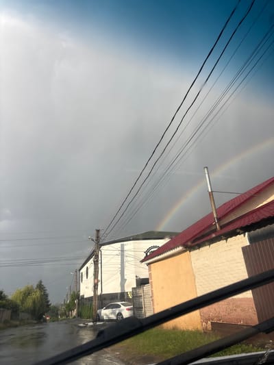 The image captures a street scene in Pokotylivka, Ukraine, following recent rainfall. The sky is a mix of light blue and heavy grey clouds, with a faint rainbow visible in the upper right quadrant. Power lines crisscross the sky. In the foreground, a wet asphalt road leads into the distance, lined with trees and fences. On the right side of the street, a white car is parked near a modern building with a white facade and dark arched windows. Adjacent to it is a smaller building with a reddish-brown roof and yellow and brick walls, featuring a metal chimney. The overall impression is one of a quiet, post-rain urban environment.