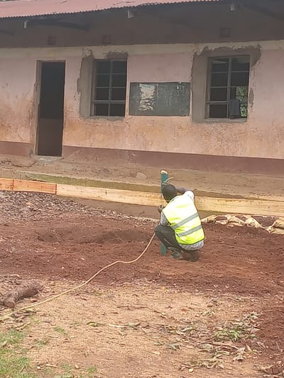A man wearing a yellow reflective safety vest is working outside a building in Idakho South ward, Kenya. The building has multiple window openings, some of which are glazed and some open. The man is bent over, seemingly engaged in work near a pile of wooden planks. The ground is a mix of soil, gravel, and sparse vegetation. An electrical cord extends away from the man on the ground.