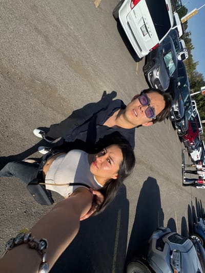 A sunlit parking lot scene features a selfie of a couple. The man, wearing glasses and a dark shirt, stands beside the woman who holds the camera. She's wearing a light top, dark jeans, and a cross-body bag. A silver charm bracelet adorns her outstretched arm. Cars line the background, including a white minivan and various SUVs. A group of people stands further back. The setting appears to be a busy outdoor area.