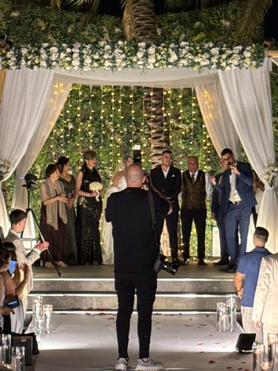 A wedding ceremony is taking place outdoors, likely in the Beer Tuvia Regional Council, Israel, at night. A chuppah adorned with white flowers and lush greenery frames the scene, with strings of warm-toned lights creating a festive backdrop. The bridal party stands on a raised platform draped with white fabric, while guests are seated on either side of a white-carpeted aisle. A photographer in black attire is positioned in the foreground, capturing the event. The groom and other male participants are dressed in suits, while the women wear elegant dresses. The atmosphere suggests a joyous celebration.
