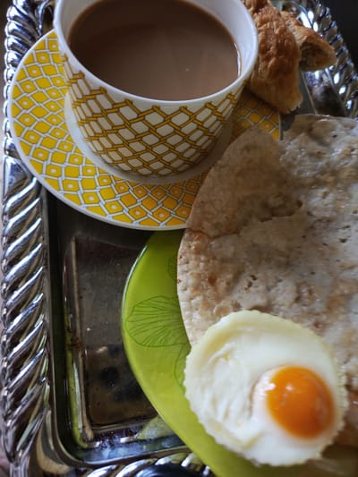 A close-up, top-down, vertical shot shows a breakfast scene on a silver-toned tray with a decorative embossed rim. The tray holds a white ceramic cup with a yellow and white geometric pattern, filled with a brown beverage, likely coffee or tea. The cup sits on a matching saucer. To the right of the cup are two pieces of light brown, flaky pastry, possibly croissants. Below the pastries and cup, on a bright green plate, is a round, flatbread, and on top of the bread is a fried egg with a bright orange yolk, cooked in a circular mold. The background is dark and out of focus. The image is captured in Alexandrovskaya, Russia.