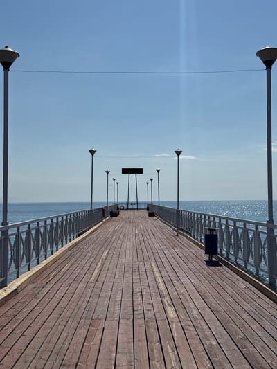 A long wooden pier extends into a calm, blue body of water under a clear sky. The pier is lined with a metal railing featuring an ornate diamond pattern. Several lampposts are spaced along the railing. At the end of the pier, a dark rectangular sign is visible, supported by green posts, with a life preserver hanging on its side. A blue trash receptacle is located on the right side of the pier. The water reflects the bright sunlight, creating a shimmering effect. The scene appears to be located in the city of Koshkol', Kyrgyzstan, likely a public recreational area by the water. There are no people visible in the image.