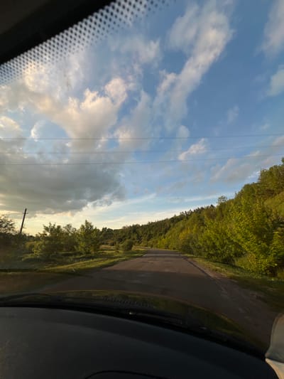 The image displays a view from inside a vehicle traveling on an asphalt road in Hrabovets, Ukraine. The scene is captured during what appears to be late afternoon or early evening, with warm sunlight illuminating the right side of the road and distant hillside. The vehicle's dark dashboard and a small portion of its hood are visible at the bottom, with the textured frit band of the windshield at the top. The upper half of the image features a bright blue sky with scattered cumulus clouds, some brightly lit and others casting shadows. Several horizontal power lines span across the sky, originating from a utility pole positioned on the left side of the road. The road itself shows signs of wear, including cracks and patches, and extends into the distance, seemingly curving to the right. The left side of the road features sparse vegetation and an open area, while the right side is bordered by a densely forested hillside with vibrant green trees and shrubs, bathed in sunlight. No individuals, animals, or other vehicles are discernible within the frame.
