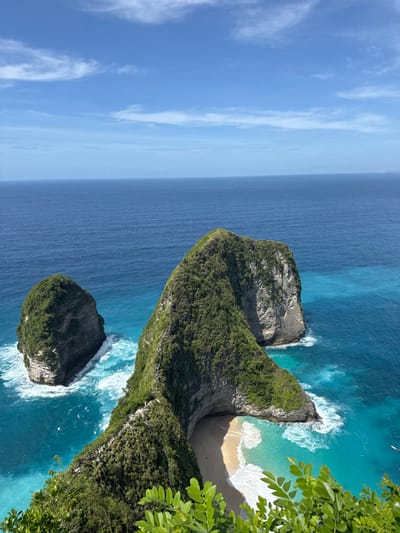 The image displays a high-angle view of a coastal landscape in Sakti, Indonesia. A prominent, irregularly shaped rocky peninsula, heavily covered with dense green vegetation, extends from the mid-left of the frame, curving rightward to form a sheltered bay. A light-colored, sheer rock face is visible on the right side of this peninsula. Within the bay, a crescent-shaped sandy beach is present, where turquoise waters meet the shore with white breaking waves. To the left of the main peninsula, a smaller, isolated, rounded rock formation, also covered in green vegetation, stands in the open ocean. The water transitions from a deep blue in the distant open sea to a lighter turquoise near the landmasses and within the bay. The sky is clear blue with sparse, wispy white clouds. Bright green foliage from a foreground plant partially obscures the lower right corner of the frame. No human figures or distinct man-made objects are observable.