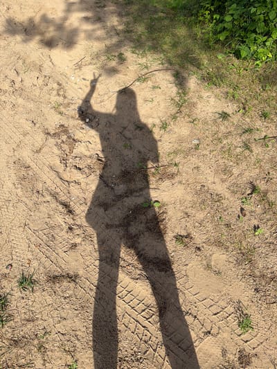 The image shows a shadow cast on sandy ground with sparse vegetation and tire tracks. The shadow appears to be of a person, with a distinct head shape, torso, and arm extended, possibly pointing. The ground is sandy and dry, with some patches of grass and pine needles. Tire tracks are visible in the sand, suggesting a vehicle has been present. The lighting indicates it is daytime, with the sun casting a long shadow. The scene is outdoors, and the presence of tire tracks and dry ground could suggest a rural or undeveloped area. There is no visible text in the image.