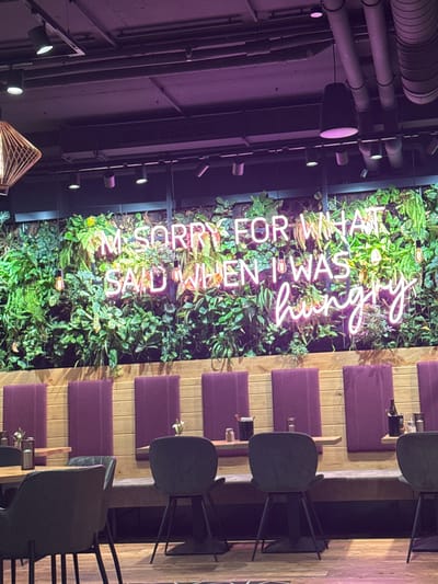 The image shows the interior of a restaurant or bar in Amstetten, Austria. A wall covered in greenery displays a neon sign reading "I'M SORRY FOR WHAT I SAID WHEN I WAS HUNGRY." Rows of tables and chairs are visible on the right side of the image, with the chairs being black and the tables having wooden surfaces and purple cushions. On the left, there are industrial-style ceiling fixtures and pipes. The overall lighting is dim and atmospheric. No people are visible.