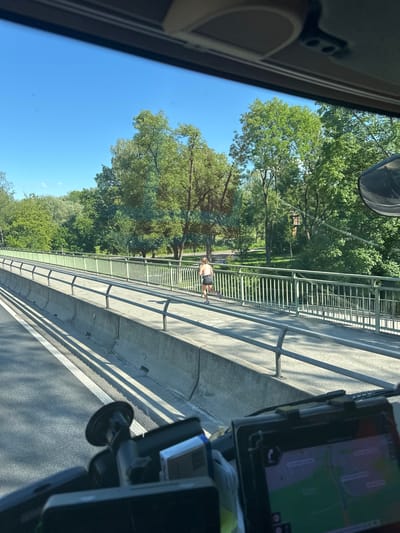 The image depicts a view from inside a vehicle, looking out at a bridge and a park on a sunny day. A person is running on the pedestrian walkway of the bridge, wearing athletic shorts and a tank top. The bridge spans over a body of water or a lower road, with lush green trees and parkland visible on the other side. The vehicle appears to be in motion, as suggested by the perspective and the light streaks on the road. The foreground shows parts of the vehicle's interior, including a dashboard with a GPS device and a suction cup mount. The overall scene suggests a typical urban or suburban environment with recreational spaces. The specific location in Augsburg, Germany, is not explicitly identifiable from the visual cues alone.