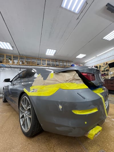 The image shows the rear passenger side of a gray BMW coupe undergoing bodywork in a garage. The car's trunk and rear bumper area are covered with masking tape and brown paper, with bright yellow painter's tape highlighting areas of repair. The garage environment is visible, with fluorescent lighting panels in the ceiling, shelving units with boxes in the background, and tools mounted on a pegboard on the right wall. The floor appears to be a speckled concrete or epoxy material. There are no people visible in the image. The car is parked, and no specific actions are taking place. The location of Veteran-2, Russia, is not ascertainable from the visual information in the image.