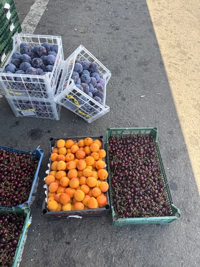 The image shows a street market scene with several crates of fruit displayed on asphalt. In the foreground, from left to right, are two blue crates filled with dark red cherries. To the right of the cherries is a black crate overflowing with ripe apricots. Behind these, in the center of the image, are two stacked white plastic crates filled with plums. The crates are arranged in a somewhat haphazard manner, suggesting a temporary market setup. The background shows a grey asphalt surface with a faint white line, possibly a road marking. A shadow falls across the scene from the right, indicating it is daytime. There are no people or animals visible in the image. The overall impression is of a fruit stand at a local market.