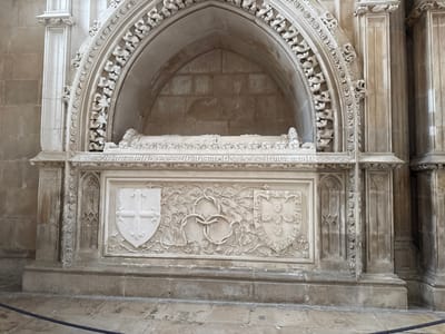 The image displays a large, intricately carved stone funerary monument located within a Gothic architectural setting in Batalha, Portugal. The monument is constructed from light-colored stone and consists of a rectangular sarcophagus base surmounted by a deeply recessed arched niche.

The front of the sarcophagus base features three distinct relief carvings. On the left, a shield displays a cross pattee with trefoil terminals. On the right, a second shield shows the Portuguese coat of arms, composed of five escutcheons each containing five bezants. Between these heraldic shields, elaborate vegetative scrollwork and a central interlaced knot design are carved. The base is flanked by decorative vertical buttresses or pilasters.

Above the sarcophagus, a horizontal frieze bears an incised inscription in an antique script. The upper section consists of a pointed arch, richly embellished with foliate and tracery patterns, framing an alcove. Within this alcove lies a recumbent stone effigy, depicting a human figure in repose, largely obscured but identifiable by its form.

The monument is integrated into a larger stone structure, characterized by Gothic-style pillars and walls made of dressed stone blocks, indicative of a monastic or ecclesiastical environment. The floor in the immediate foreground is paved with stone. A dark rope barrier is visible horizontally across the lower part of the frame, marking a designated viewing area. No individuals or active subjects are present.