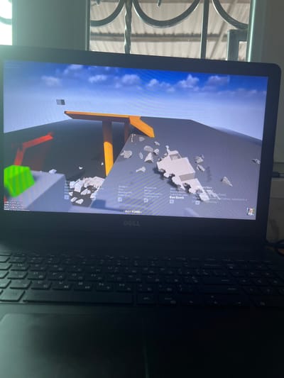 The media file shows a close-up of a laptop screen displaying a video game. The game appears to be a physics-based sandbox game with a 3D environment. Debris, possibly from an explosion, is scattered across the screen. A menu with various items like "Pipe bomb," "Shotgun," and "Bay Bomb" is visible on the right side of the screen, along with controls like "LMB to plant bomb" and "RMB to detonate." The game's aesthetic is blocky and somewhat simplistic. The laptop itself is black and the keyboard is partially visible, showing Cyrillic and Latin characters. In the background, through a window or opening, a glimpse of what appears to be a corrugated metal roof and some light-colored structures can be seen, suggesting an outdoor or industrial setting. The lighting on the screen suggests it's daytime. No people are directly visible in the image, but the content on the screen points to a user interacting with the game. The text visible on the screen is a mix of English and Russian, with game-specific instructions and a snippet of Russian text that translates to "To activate Windows, go to 'Settings'." The overall scene is dominated by the bright display of the laptop screen.