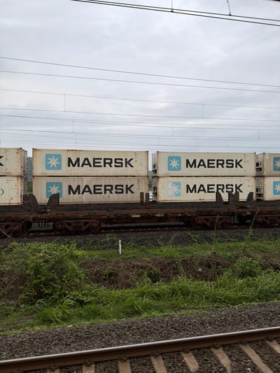 The image shows a train with multiple Maersk shipping containers stacked two high on flatbed railcars. The train is on tracks surrounded by lush green vegetation and some rocky terrain. Overhead power lines are visible against a cloudy sky. The scene is likely near Saphale, India, given the presence of a train line and the surrounding landscape. There are no people visible in the image, and the train is stationary.