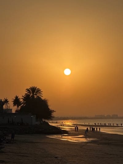 The scene captures a sunset over a beach in Bawshar Province, Oman. The upper portion of the frame is filled with an orange-yellow sky, featuring a bright, circular white sun positioned in the upper-middle right. The foreground consists of dark sandy beach. Along the left edge, multiple silhouetted palm trees and dense vegetation flank a single-story building equipped with outdoor seating and umbrellas. Construction scaffolding is visible on the far left, near one of the palm trees. Numerous individuals, mostly silhouetted, are present on the beach and wading at the water's edge. A distinct group walks along the shoreline in the midground, where the shallow water reflects the setting sun in a shimmering golden path. One individual in the midground appears to be kicking a light-colored object near the water. In the distant background, a hazy, dark silhouette of land or a city skyline is visible on the horizon.