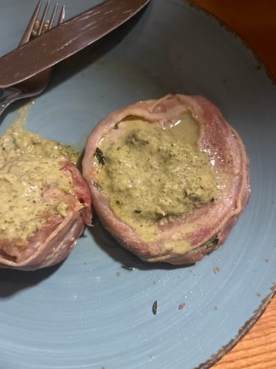 This close-up image presents two portions of a prepared meal served on a light blue ceramic plate. Each portion consists of a core of meat, likely chicken or pork, wrapped in thinly sliced cured meat such as bacon or prosciutto. The center of each wrapped item is filled with a generous amount of a thick, pale green, creamy sauce, which appears to be herb-based or pesto-like.
The upper portion of the dish is neatly presented, showcasing the sauce contained within the meat wrap. The lower portion has some of the green sauce spilled onto the plate, suggesting it may have been partially cut or disturbed. The plate itself features a slightly textured, rustic rim. A silver knife with a serrated blade and a matching fork are placed on the left side of the plate, indicating the meal is ready to be eaten.
The scene is set indoors, likely at a dining table, with a warm, wooden surface visible in the background. The lighting is relatively dim, suggesting an evening meal or an intimate indoor setting. There are no individuals or visible text in the image. The overall impression is of a gourmet or thoughtfully prepared dish.