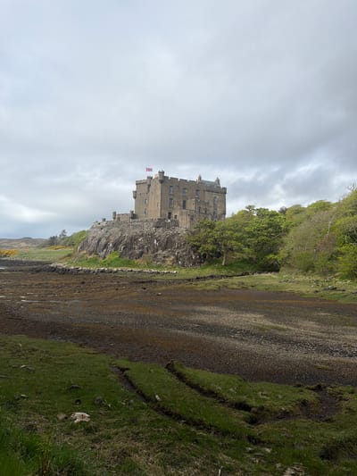 The image depicts Dunvegan Castle in Dunvegan, United Kingdom, positioned atop a rocky outcrop on the coastline. The castle, constructed of gray stone, features multiple stories, towers, and windows. Two flags are visible atop the castle. The sky is overcast with gray clouds. The foreground consists of a rocky shoreline with seaweed and green grass. A strip of land covered in rocks, dirt and grass is also visible. The perspective of the image is landscape view but tilted to be in portrait position.