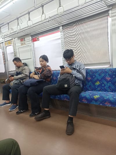 Three individuals are seated on blue and white patterned train seats facing the right side of the image. The man on the right is wearing a light blue and white striped shirt, blue jeans, a black backpack, and black shoes. He is looking down at his phone in his right hand. The woman in the middle is wearing a brown and white patterned blouse, black pants, and black shoes. She is looking at her phone in her left hand. The man on the left is wearing a blue jacket, dark blue jeans, and black shoes. He is looking down at his phone in his right hand. They are all on a train in Bojong Gede, Indonesia (latitude: -6.501, longitude: 106.796). The interior of the train is lined with a light grey material and decorated with silver metal handles. There are two windows in the background. The train appears to be empty besides these three individuals.