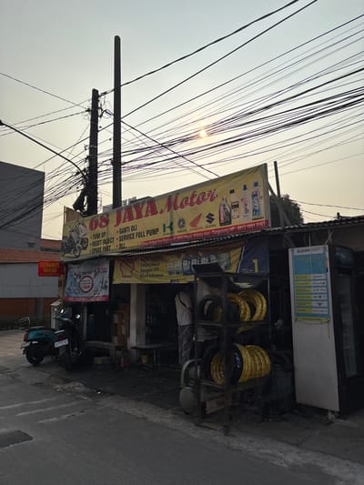 The image depicts the storefront of "68 JAYA MOTOR," a motorcycle repair shop located at Jl. Danau Kelapa Dua Raya No. 97, Kelapa Dua, Tangerang, Indonesia. The shop specializes in oil changes, full-service pump repairs, and spare parts for motorcycles, as indicated by the yellow banner. Several motorcycles are visible, including a teal scooter parked on the left and other bikes inside the shop. Tires are stacked on display racks in front of the shop. A man in a white shirt and checkered pants is seen standing inside the workshop. The sky is overcast with a hazy sun peeking through a dense network of electrical wires overhead. The overall environment appears to be a street in an urban or suburban area.