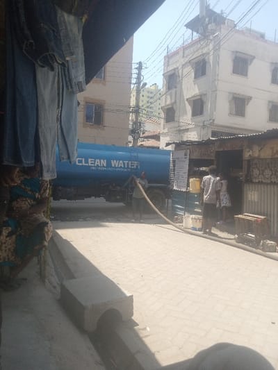 This image depicts a daytime street scene in Tudor ward, Kenya, characterized by urban infrastructure and activity. In the immediate foreground on the left, several items of blue and light blue denim clothing hang from an unseen structure, partially obscuring a seated individual wearing a patterned blue and brown dress. The ground consists of light-colored interlocking pavers, with a concrete drainage channel running along the bottom left, partially covered by a precast concrete section.
Centered in the midground is a large, blue tanker truck marked with white text "CLEAN WATER" and two phone numbers: "CALL: 0720427079 - 071249210". A male individual, dressed in a light-colored shirt and dark shorts, stands beside the truck, actively managing a large, coiled beige hose extending from the tanker. To the right of the tanker, a small commercial structure with corrugated metal walls is visible, displaying a white informational poster. In front of this structure, a male individual in a light shirt and dark shorts stands with his back to the viewer, facing a female in a white patterned dress and a smaller individual, all appearing to be near the shop's entrance. Several white buckets and a yellow jerry can are positioned on the ground near the shop. A wooden crate or pallet rests on the ground to the far right.
The background features several multi-story buildings. Directly behind the tanker, a beige-colored building with multiple windows is visible. To its right, a taller, white, five-story building with numerous dark-framed windows and a flat roof with utility structures is present. Further in the distance, a light green or yellow high-rise building can be seen. A dense network of overhead utility wires and poles traverses the clear, bright sky.