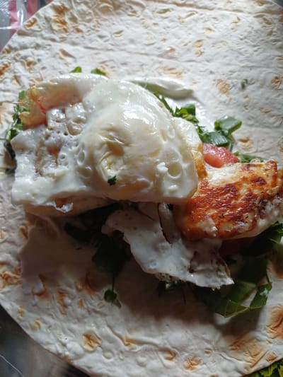 A close-up, overhead shot shows a partially wrapped lavash (flatbread) filled with ingredients, resting on a metallic surface. The lavash is white with lightly browned blistered spots. Inside the lavash, there are fried eggs with visible whites and yolks, pieces of grilled halloumi cheese with a golden-brown crust, chopped green onions, and sliced red tomatoes. A white sauce, likely mayonnaise or yogurt-based, is drizzled over the fillings. The scene does not contain any recognizable landmarks or context to indicate a specific location in Sulymivka, Ukraine.