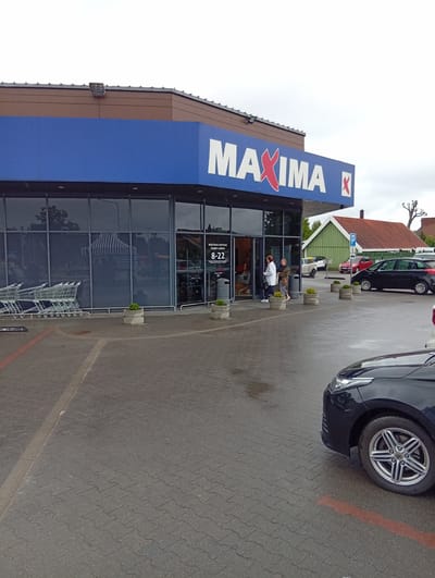 The image shows the entrance of a "Maxima" supermarket in Kuršėnai, Lithuania. Two women are walking out of the store. The supermarket has a blue facade with the store's name in white letters and red "X" logos. In front of the store are rows of shopping carts and planters with small green plants. The ground is wet, suggesting recent rain. Several cars are parked in the lot, including a black car in the foreground. A green building and a red-roofed house are visible behind the supermarket. The sky is overcast.