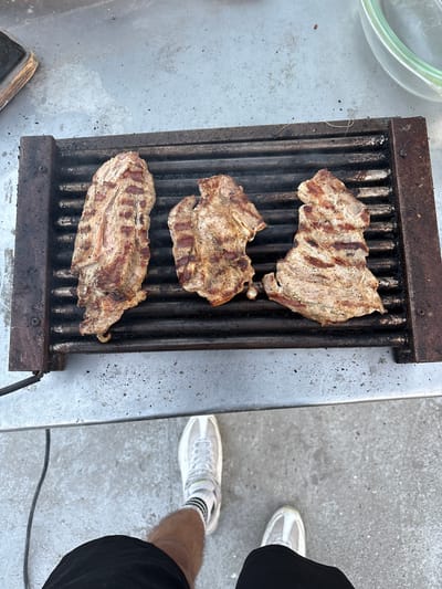 The image shows three pieces of grilled meat, likely steak, on a grill. The grill is placed on a stainless steel surface. In the upper left corner, part of a glass bowl is visible. To the right, a person is standing wearing white sneakers, black shorts, and socks with black stripes. The presence of a power cable suggests the grill might be electric. No information suggests the image was taken in Butovo, Bulgaria.