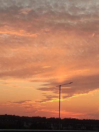 The image shows a sunset sky over Kraskovo, Russia, with a silhouetted street lamp in the foreground. The sky is filled with clouds that are illuminated by the setting sun, casting warm orange and pink hues across the scene. Below the sky, a dark line of trees and buildings forms the horizon, indicating a residential area. The overall impression is one of natural beauty and peacefulness.