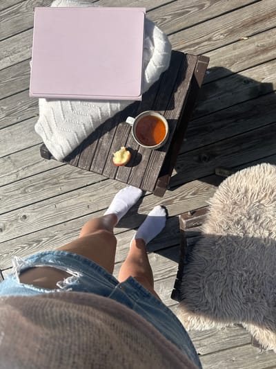 This is a high-angle, full-shot, outdoor photograph taken on a wooden deck. The scene depicts a person sitting on a chair with a fluffy sheepskin rug. In the foreground, the legs of the person are visible, wearing white socks and ripped denim shorts, with a light-colored sweater or shawl draped over their lap.

On a low wooden table, to the left of the person, are several items. A light pink tablet rests on a white knitted blanket. Next to the tablet is a mug filled with an amber-colored liquid, likely tea or coffee, and a partially eaten piece of apple. The table is casting a shadow on the wooden deck.

The environment appears to be a residential outdoor space, possibly a patio or balcony. The presence of the tablet, mug, and fruit suggests a moment of relaxation or work outdoors. The sunlight is casting strong shadows, indicating a sunny day.

There is no specific indication of the location being in the city of Znamenka, Russia, in the provided image. The scene is general and could be in many locations.