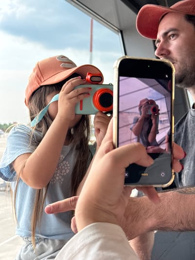 In a sunlit interior, likely an airport terminal given the large window and sky visible, a young girl wearing an orange cap and a light blue t-shirt is focused on taking a photograph with a teal and orange toy camera. Her hands are positioned to operate the camera, and a turquoise strap hangs from it. To her right, a man wearing a red cap and a dark t-shirt is looking towards the girl, with his arm held out holding a gold-rimmed smartphone. The phone's screen displays a reflection of the girl taking a picture with her toy camera, creating a layered photographic moment. The environment outside the window appears to be an airfield or open area with a cloudy sky. The perspective suggests the photo is taken from a third person's viewpoint, possibly a parent, capturing this interaction. No specific landmarks identify the location as Bol'shie Kabany, Russia.