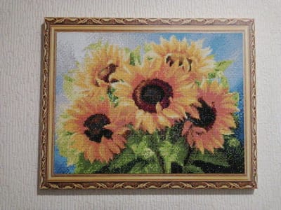 The image shows a framed diamond painting of sunflowers, hanging on a textured, off-white wall. The painting depicts a cluster of sunflowers with yellow and orange petals, dark brown centers, and green leaves against a blue background. The diamond painting technique creates a mosaic-like effect with small, faceted beads. The frame is ornate, with a gold and brown patterned border. The wall appears to be an interior wall, possibly in a residential setting. There are no people or animals visible in the image. The location is stated to be in the settlement of the experimental farm "Ermolino," Russia.