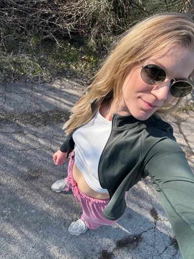 A blonde female is captured in a high-angle outdoor selfie, standing on a cracked asphalt or concrete surface in the city of Dolni Rakovets, Bulgaria. She wears gold-rimmed oval sunglasses, the right lens displaying a hairline crack and a visible "Ray-Ban" logo. Her attire consists of a dark green long-sleeved zip-up jacket, partially unzipped over a white ribbed cropped tank top that exposes her midriff. Below, she wears pink drawstring sweatpants and light grey athletic shoes. Her right arm is extended to operate the camera, and she displays a slight smile. A dark spot is present on her right cheekbone. The background shows sparse vegetation with bare branches and green patches, consistent with an outdoor, possibly roadside or path, environment. Strong shadows are cast on the ground, indicating direct sunlight.