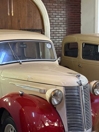 The image depicts two vintage automobiles displayed indoors at a location in Ramboda, Sri Lanka, likely the Ramboda Falls Hotel. In the foreground, a two-tone classic car is prominent, featuring a cream-colored body and hood contrasting with glossy red front fenders. Its front fascia includes a chrome vertical grille, a chrome-winged hood ornament, and two circular headlights with chrome surrounds. A single windshield wiper is positioned on the driver's side of the windshield, which also has a circular white sticker on its passenger side. A chrome trim piece extends horizontally along the side of the hood, and a small, red-capped signal light is mounted on the apex of the left front fender. Part of a white wheel rim is visible beneath the fender. Behind and to the right of this vehicle, a second, fully cream-colored vintage car is partially visible, showing its side profile including a window and part of its roofline. The background features a wall constructed of reddish-brown bricks, and to the upper left, a large arched wooden doorway framed by a light-colored archway. No individuals are present in the scene.