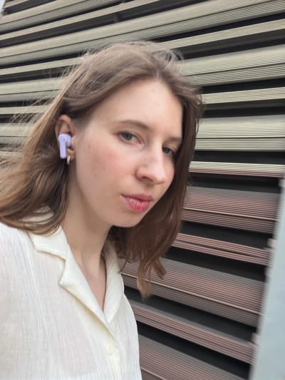 A young woman with long brown hair, wearing a light-colored collared shirt and a lavender earbud, is captured in a close-up portrait against a background of horizontal striped panels. She is looking towards the camera with her mouth slightly parted. The scene is set outdoors, suggesting an urban environment, potentially within Pocheon-si, South Korea.