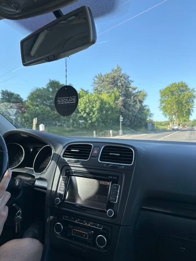 The image is taken from inside a car, looking out through the windshield. A person's hand is visible on the steering wheel, and the car's dashboard with the radio and air conditioning controls is in the foreground. A black car air freshener hangs from the rearview mirror, displaying the text "VERGNON AUTO GROUPE". Outside the car, a tree-lined road is visible under a clear blue sky. The scene appears to be a sunny day in a rural or suburban area. The exact location within Bagard, France, cannot be determined from the visual information alone.