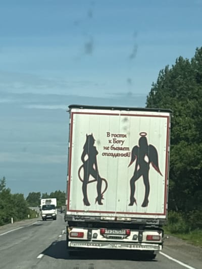 This image captures the rear of a white truck trailer driving on a paved road. The trailer's back features a graphic of two silhouettes of women. The silhouette on the left has horns and a tail, resembling a devil. The silhouette on the right has wings and a halo, representing an angel. Above the silhouettes, in Russian text, is written "В гости к Богу не бывает опозданий!" which translates to "You are never late for God!". The road is bordered by green trees and under a bright blue sky with some clouds. In the distance, another white truck is visible. The license plate of the truck in the foreground is partially visible and appears to be Russian. The image is taken from a low angle, suggesting the photo was taken from another vehicle.