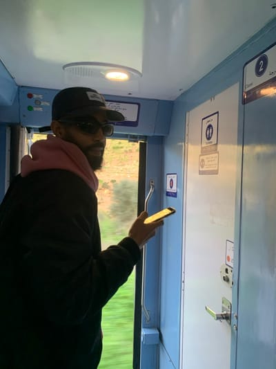 The image depicts the interior of a moving train or bus, seen from a low angle. In the foreground, a person is partially visible, primarily their upper body, facing towards the right side of the frame. This individual appears to be a man with a short beard, wearing dark sunglasses, a black baseball cap, a dark-colored jacket, and a light pink hooded garment (possibly a scarf or hoodie) underneath. His right arm is raised, and he is holding a smartphone, the screen of which is brightly illuminated by direct sunlight, casting a reflection.

The interior of the vehicle features light blue and white panels on the walls and ceiling. On the left side, embedded in a white panel, is a circular light fixture emitting a warm, orange-yellow glow. Above the window area, several signs are affixed to the white paneling. One prominent sign displays two stick figures (representing male and female) within a blue square, accompanied by the word "Toilettes" (French for toilets) and Arabic script above it. To the left, another sign shows the number "2" inside a blue circle, with Arabic text above and "Seconde" (French for second) below, likely indicating a class or carriage number. Further to the right, a silver handle or latch mechanism is visible on an overhead panel. Below the "Toilettes" sign, a smaller, circular red sign with an indistinguishable symbol is present. A horizontal silver grab bar or handle runs along the window line. Another white panel with Arabic script is visible on the lower left side.

Through the window on the right, a blurry, vibrant green landscape can be seen, indicating the vehicle is in motion and traveling through a vegetated area. Strong natural sunlight streams in from outside, highlighting the person's hand and phone, and creating bright reflections on the interior surfaces. The overall impression is one of travel during daylight hours, with the strong sunlight suggesting morning or afternoon.