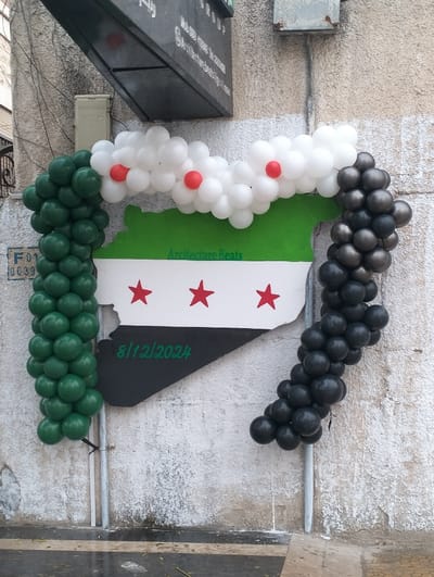 A celebration display showcasing the Syrian flag colors. Green and black balloons adorn the sides. A representation of the country's shape is painted in green, white, and black. White balloons with red accents run across the top. Three red stars are in the white stripe. The date 8/12/2024 is written in green on the black section of the flag. The display is attached to a weathered wall with metal poles.
