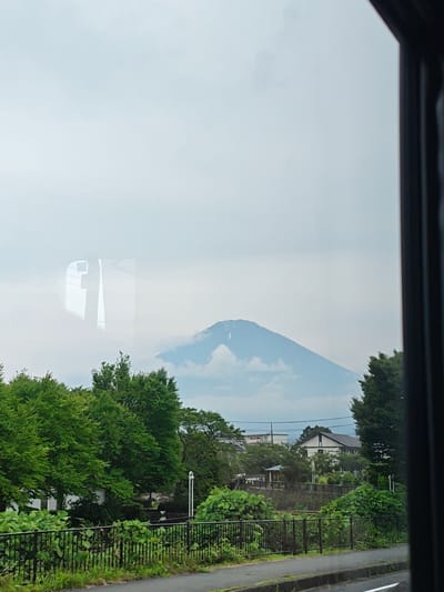 The image depicts a view of Mount Fuji, partially obscured by clouds, from the city of Gotemba, Japan. The foreground features lush green trees lining a black metal fence, with a paved road visible beyond. In the mid-ground, residential buildings and more trees are scattered. The sky is overcast, contributing to the soft lighting of the scene. The reflection of what appears to be a window frame is visible on the mountain itself.