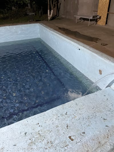 The image shows an outdoor swimming pool at night, with water flowing into it from a pipe on the right side. The pool walls are white and appear to be made of concrete. The bottom of the pool is tiled with a dark blue pattern. In the background, there is a concrete area with a table and chairs, and some trees and vegetation. The overall environment appears to be somewhat neglected. The location is identified as Mashat, Kazakhstan.