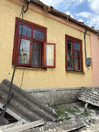 The image displays the exterior of a damaged single-story residential building in Nedryhailiv, Ukraine, under a partially cloudy blue sky. The building's facade is textured light yellow stucco, with a section of light pink stucco visible on the right. A gray concrete base with a faux-stone pattern lines the lower portion of the wall. Two windows with dark red frames are present. The left window features multiple panes, with its lower-right pane open outwards, revealing a textured glass insert. The right window's glass is extensively shattered and partially missing, with fragments still attached to the frame. Curtains are visible behind both windows.

Above the windows, the roof eaves show significant damage, including exposed and splintered wood and torn material. A loose, dark electrical cable hangs from the upper left of the building. Another black electrical conduit runs vertically on the right side, adjacent to an external electrical meter box mounted on the wall. The immediate foreground is covered with multiple large, broken pieces of dark gray corrugated roofing material, scattered across patchy green grass and dirt. White, fibrous debris is intermingled with the roofing fragments and ground cover. No individuals are visible.