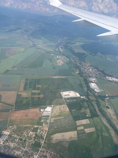 An aerial view captures a segment of the landscape around Grigorevo, Bulgaria. The white wing of an aircraft occupies the upper right quadrant, featuring visible flap fairings on its underside. The scene below comprises a mosaic of agricultural fields in varied shades of green and brown, interspersed with areas of dense woodland. Distant, forested mountains define the horizon under a partially cloudy sky.

Multiple residential clusters are evident; a compact village with numerous houses featuring orange-red roofs is centrally located in the upper-middle section, traversed by local roads. Another denser residential zone is visible in the lower-left corner. A prominent multi-lane highway, including an interchange, carves through the landscape in the upper-middle area. Additional roads and tracks connect fields and settlements. A linear feature consistent with a railway line runs horizontally across the mid-section. Large, light-roofed industrial or commercial buildings, some with rooftop solar panels, are positioned near roads in the mid-left and mid-right sections of the image.