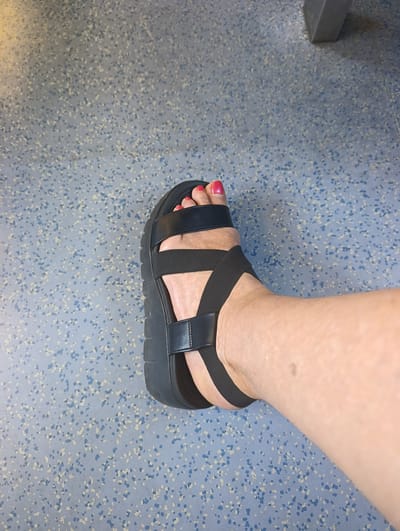 A person's right foot, adorned with bright pink nail polish on the toenails, is shown wearing a black platform sandal. The sandal features two thick black straps across the foot, one of which crosses over the other. The sole of the sandal is a substantial black platform with a ridged texture. The foot and sandal are positioned on a speckled blue and grey floor, which appears to be linoleum or a similar material. The background is blurred, suggesting the image was taken in a public space, potentially a transit area, as indicated by a glimpse of a grey, angular structure that might be part of a seat or column. The context of Palmela, Portugal, is not discernible from the visual information provided in the image.