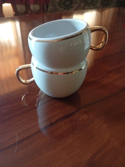 Two white ceramic cups with gold trim are stacked on top of each other. The cups are sitting on a dark wooden table. The background is blurred but appears to be an indoor setting with some furniture and drapery. The lighting suggests it is daytime.