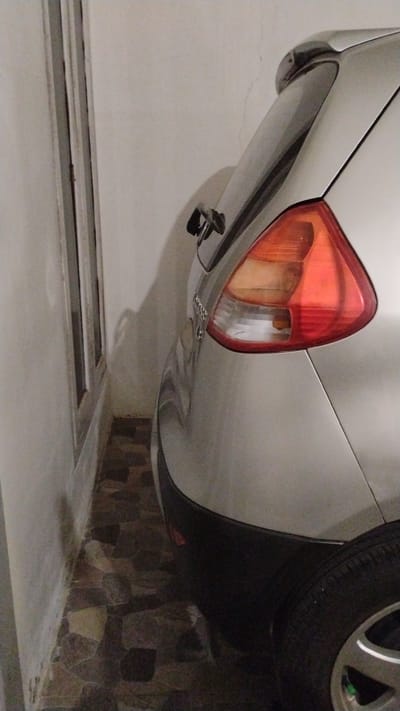 A silver car is parked in a narrow space between a white wall and a window frame. The car's rear taillight, bumper, and tire are visible. The ground is covered with irregularly shaped stone tiles. The scene appears to be an outdoor parking area, possibly in a residential or commercial setting, in Banjarsari, Indonesia.
