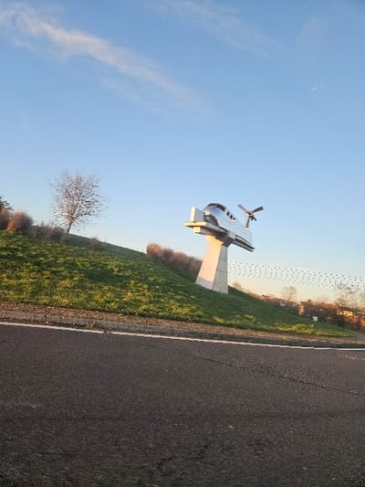 An outdoor scene in Gometz-la-Ville, France, is captured under a clear blue sky with scattered wispy clouds. The foreground presents an asphalt road displaying a solid white edge line and minor surface cracks, leading to a concrete curb. Beyond the curb, a steep, green grassy slope ascends. On this slope, a bare deciduous tree stands to the left. Further right, a prominent monument, depicting a streamlined metallic vehicle resembling an aerotrain or high-speed train with a cockpit-like front and rear fins, is erected on a tall, angled concrete pylon anchored into the slope. In the distant background, a line of trees and indistinct building structures are visible. A flock of numerous small, dark birds flies in a horizontal formation across the sky. Natural sunlight illuminates the scene from the right, casting soft shadows. No human presence or active interactions are observed.