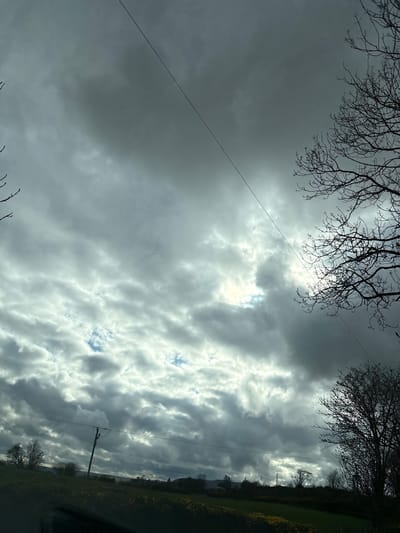 The image captures a cloudy sky with bare tree branches in the upper corners. A wire stretches diagonally across the left side of the frame. In the distance, a green field and a utility pole are visible on the right. The photo appears to have been taken in or near Stiperstones, United Kingdom, given the landscape and cloud formations.