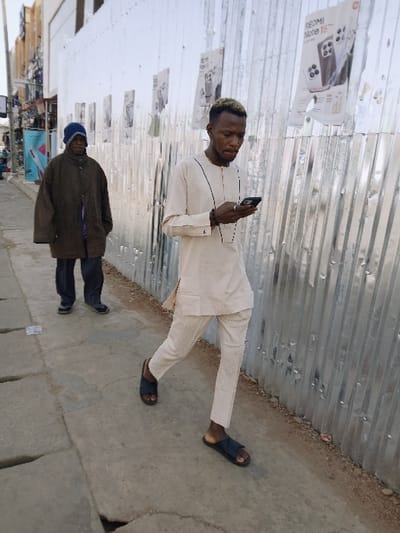The image depicts an urban outdoor scene on a concrete sidewalk in the city of Kungulu School, Zambia. Two males are present. In the foreground, a young adult male with light-tipped hair walks towards the left, actively engaged with a dark-colored smartphone held in both hands. He wears a light beige long-sleeved tunic with black piping, matching trousers, and dark open-toe sandals. A dark bracelet is visible on his left wrist and a ring on his right ring finger. Behind him, an older adult male, wearing a dark blue beanie, an oversized dark brown coat, dark trousers, and dark closed-toe shoes, walks in the same direction. He appears to be observing the foreground. To the immediate right of the subjects, a corrugated metal wall, possibly a temporary construction barrier, is visible. Several white posters are affixed to this wall, one clearly advertising "REDMI Note 15" with a smartphone image. The concrete sidewalk exhibits visible cracks. In the distant left background, obscured structures and overhead wires are discernible under bright daylight conditions. There is no direct interaction between the two individuals.