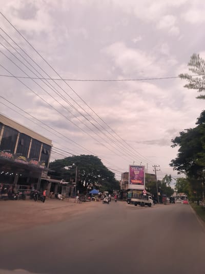 This image captures a bustling street scene in Lambaro, Indonesia, under an overcast sky, suggesting either early morning or late afternoon light. Numerous dark power lines stretch across the upper part of the frame, contrasting with the light pinkish-grey clouds.
On the left, a multi-story building with a light-colored facade and dark windows features a ground-floor retail space with awnings displaying "MW" branding. Several motorcycles are parked in front, and people are visible near the storefront. Behind this building, a large, dense green tree stands prominently.
Towards the right side of the road, a large billboard is visible, advertising "VEEVA GRAPE" for "Rp 35.000/box" and displaying a "21+" age restriction symbol. A white pickup truck is parked beneath the billboard. Utility poles with additional power lines are scattered along this side of the street. Further down, more buildings and vehicles, including cars, can be seen in the distance. A red circular sign with an "R" is also noticeable near the billboard.
The road itself appears to have both paved and unpaved sections, with dirt shoulders on the left. The scene is active, with people walking, riding motorcycles, and engaging in daily activities. A blue umbrella near the center indicates a roadside vendor or stall. The overall impression is that of an active commercial and residential street.
