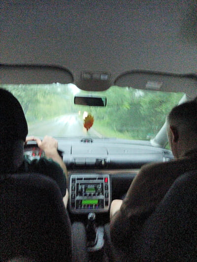 The image captures the interior of a car from a rear-facing perspective, showing two individuals. The person in the driver's seat, on the left, is seen from the back of their head and shoulder, with their hands on the steering wheel. Their body is slightly turned, and their arm is extended. To the right, a second person is visible from the back of their head and shoulder, facing forward.
The scene inside the car includes the dashboard, with a radio or infotainment system displaying green text, and the gear shift. A car air freshener, shaped like a leaf, hangs from the rearview mirror.
Outside the car, visible through the windshield, is a wet road, indicating rain. The surrounding environment appears to be a rural or wooded area with lush green trees. The lighting suggests it is daytime, likely overcast due to the weather.
The image conveys a sense of travel, with the individuals in the car engaged in the activity of driving. The grainy quality of the image, possibly due to low light or camera settings, adds to a somewhat indistinct and atmospheric feel. The location context provided is Šternov, Czechia.