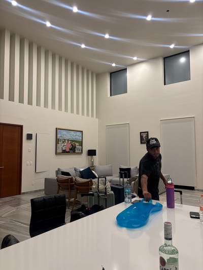 A man stands at a white countertop in a modern interior in Santiago Etla, Mexico. He wears a black t-shirt and black cap, and he holds a blue plastic bag in his hands. On the counter are a purple water bottle, a partially empty clear glass bottle, and other items. Behind the man is a living room area with a grey sofa, chairs, and a glass coffee table. A framed painting hangs on the wall. In the background, a staircase with horizontal white bars is visible. Above the staircase, there are two square windows and a row of recessed lights on the wall. The room is well-lit, indicating the presence of electrical lighting fixtures.